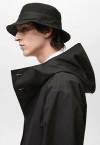 Pălărie neagră tip bucket cu margine lată, purtată cu o jacheta neagră cu glugă, având fermoar și închidere cu capse. Material neted și design minimal.
