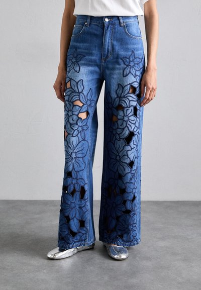 Jeans in denim blu a vita alta con ampie ricami floreali e design a taglio. Forma svasata con orli grezzi e tasche anteriori. Scarpe argento.