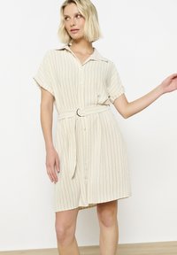 Beige napakka mekko, jossa on lyhyet hihat, pystysuorat mustat raidat, vyö pyöreällä soljella vyötäröllä ja rento istuvuus.