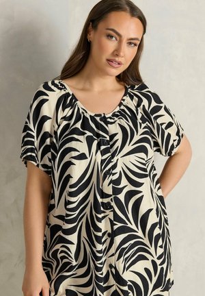 Frau mit langen braunen Haaren, die ein schwarz-weiß gemustertes Hemd mit Knöpfen trägt und vor einem neutralen Hintergrund steht.