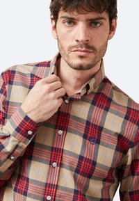 Camisa de cuadros en beige, rojo y azul marino con cuello abotonado, que presenta bordado personalizado y botones en contraste. Tejido texturizado.