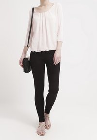 Helles rosa, locker sitzendes Blusenshirt mit Falten; schwarze, figurbetonte Leggings; schwarze Umhängetasche; braune Sandalen mit dekorativen Riemen.