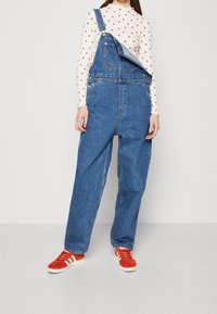 Salopette in denim di colore azzurro chiaro con una vestibilità ampia, dotata di bretelle regolabili e tasche frontali, abbinata a scarpe da ginnastica rosse.