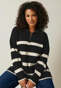 Schwarz-weiß gestreifter Pullover mit einem halben Reißverschluss-Kragen, weiten Ärmeln und einer weichen Textur, kombiniert mit dunkelblauen Jeans.