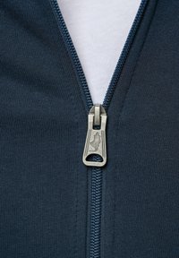 Gros plan sur une fermeture éclair en tissu bleu marine partiellement fermée avec une tirette en métal présentant un petit logo en silhouette.