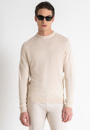 Maglione lungo a maniche lunghe beige chiaro con scollo rotondo, realizzato in morbido tessuto a maglia. Modello che indossa occhiali da sole neri e pantaloni chiari.