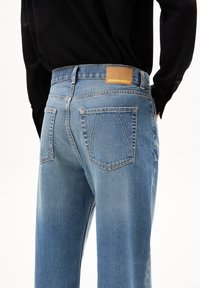 Jeans de mezclilla azul claro con cintura alta, que presentan dos bolsillos traseros y una etiqueta de cuero marrón en la cinturilla. La tela texturizada muestra un ligero desvanecimiento.