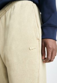 Beige sporta bikses, kas izgatavotas no mīksta auduma, ar sānu kabatām un izšūtu logotipu kreisajā augšstilbā. Roku atpūta uz jostas.