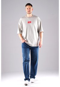 Helles graues, übergroßes T-Shirt mit einem roten Logo-Patch, kombiniert mit blauen geraden Jeans und weißen Sneakern. Schlichter Hintergrund.