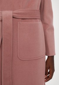 Mjuk, rosa ullblandad kappa med bälte, bred krage och stor ficka. Tyget har en slät, texturerad yta.