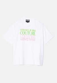 Bílá bavlněná tričko s oversized textem v zelené a růžové barvě, který říká "Versace Jeans Couture" v moderním písmu. Krátké rukávy, kulatý výstřih.