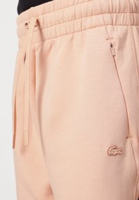 Light pink sweatpants with an elastic waistband, drawstring, and side zipper pockets. Features a small embroidered logo. Soft texture.