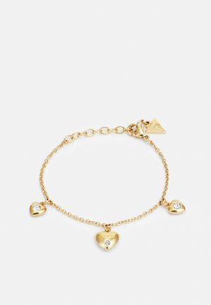 Bracelet délicat en chaîne d’or avec trois breloques en forme de cœur, chacune ornée d’une petite pierre transparente, et une étiquette de marque triangulaire près du fermoir.