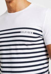 Muž na sobě má bílé tričko s horizontálními námořnickými pruhy a na hrudi je velkými písmeny napsáno "HILFIGER".