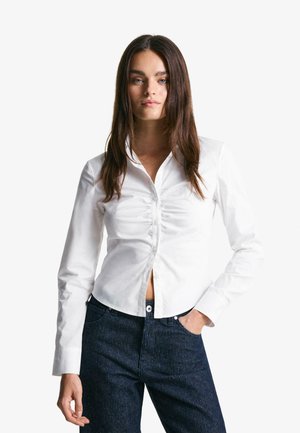 Camicia bianca con bottoni e maniche lunghe, con tessuto arricciato sul davanti. Abbinata a pantaloni in denim scuro, con righe verticali.