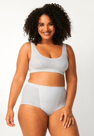 Femme souriante aux cheveux bouclés portant un soutien-gorge sans fil gris clair et une culotte taille haute, posant devant un fond blanc uni.