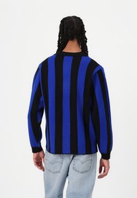 Strikkegenser med kraftige vertikale svarte og blå striper, lange ermer, ribbestrikkede mansjetter og en avslappet passform. Brukt sammen med lyseblå jeans.