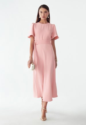 Woman wearing a light pink belted midi dress with flutter sleeves, holding a small silver clutch, and wearing gold high-heeled sandals.