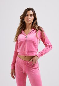 Rózsaszín velúr melegítő szett, amely egy rövidített cipzáras kapucnis pulcsiból áll elülső zsebbel, valamint hozzá illő, húzózsinóros nadrágból. Hímzett logó részlet.