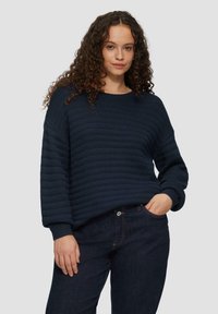 Marineblauer Rippenstrickpullover mit Rundhalsausschnitt und langen Ärmeln, im lässigen Schnitt und mit strukturiertem Muster. Getragen mit dunklen Jeans.
