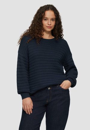 Granatowy sweter z prążkowanego materiału, z okrągłym dekoltem i długimi rękawami, charakteryzujący się luźnym krojem i teksturowanym wzorem. Noszony z ciemnymi dżinsami.