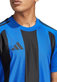 Blaues und schwarzes Sporttrikot mit Rundhalsausschnitt, das drei schwarze Streifen auf den Schultern und ein stilisiertes schwarzes Logo auf der Brust aufweist.