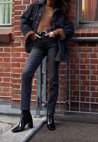 Chaqueta de mezclilla negra y cuello alto a rayas, combinada con botines negros de tacón alto, jeans oscuros, sobre un fondo de ladrillo.