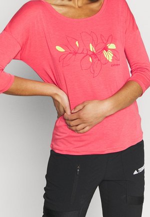 Persoon draagt een koraalroze longsleeve met bloemenprint en zwarte Adidas Terrex broek, handen in elkaar gevouwen ter hoogte van de taille.