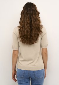 Haut en tricot beige avec de courtes manches bouffantes, tissu texturé et ourlet ajusté, associé à un jean bleu clair, vu de dos.