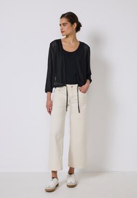 Czarna siateczkowa kurtka ze skróconym krojem, luźny fason, otwarty przód ze sznurkiem, noszona na czarnym topie i kremowych szerokich spodniach, zestawiona z białymi trampkami.