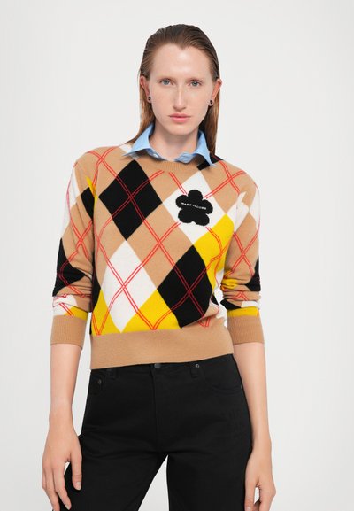 Knit sweater with argyle pattern in beige, black, yellow, and white; features red lines and a black emblem with "Marc Jacobs." Black trousers.
