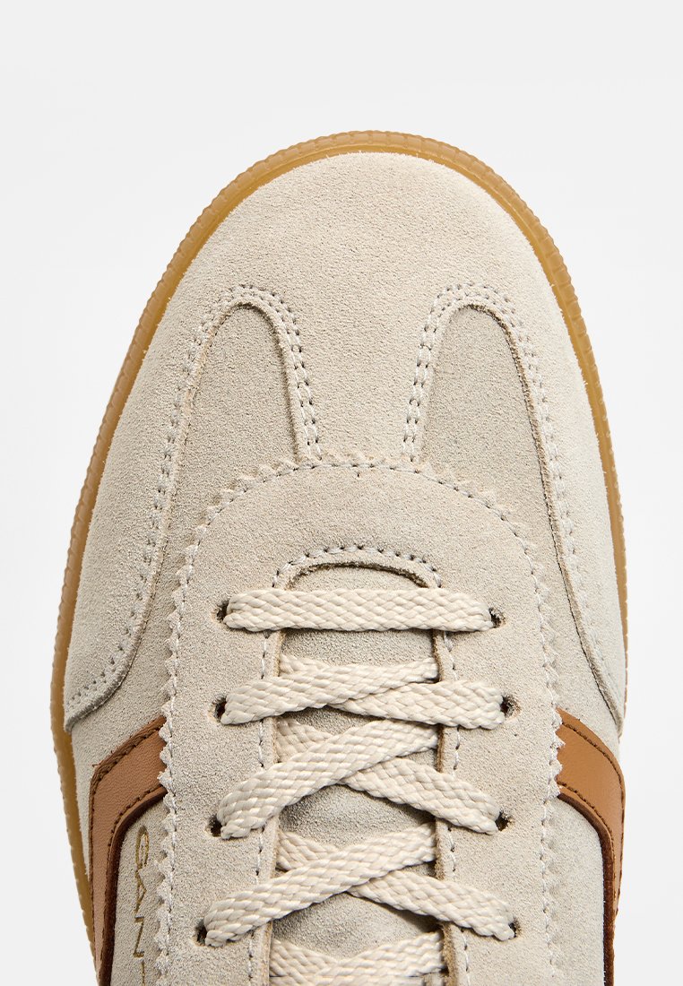 Zapatillas de ante en beige claro con suela de goma. Presenta agujetas en crema y un acento de cuero marrón en el costado. Diseño clásico.