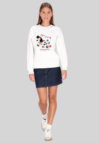 Sudadera blanca con un gráfico colorido de cachorros y texto, combinada con una falda de mezclilla. La modelo lleva zapatillas blancas con detalles marrones.
