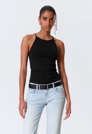 Femme aux cheveux bouclés portant un haut noir sans manches et un jean bleu clair avec une ceinture noire, debout devant un fond uni.