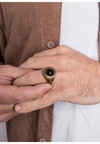 Anello color oro con un centro in pietra nera, con dettagli intricati e una fascia texturizzata, indossato su una mano con evidente peluria.