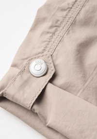 Tessuto beige con una texture liscia e cuciture visibili, caratterizzato da un bottone argentato con scanalature e dettagli del marchio in rilievo.