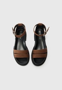 Sandales en cuir synthétique brun avec deux larges sangles et une sangle de cheville réglable, dotées d'une semelle en caoutchouc noir texturé.
