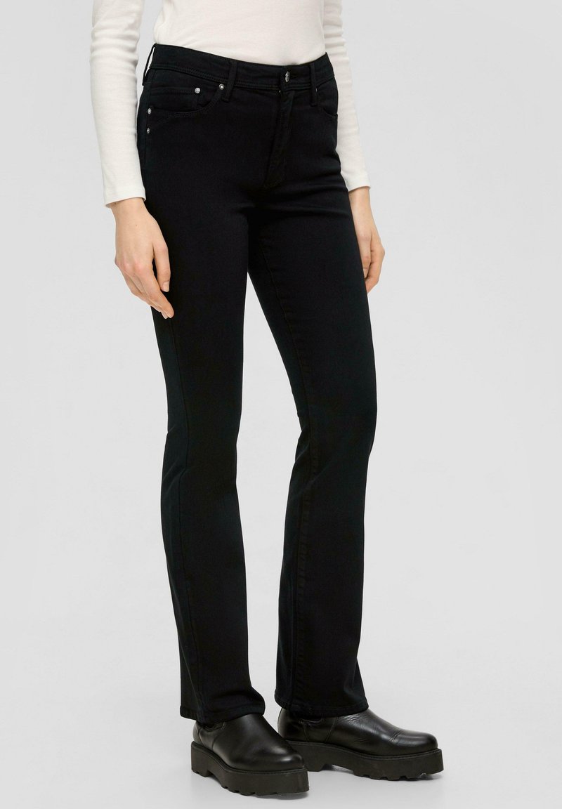 Frau in schwarzen Slim-Fit-Jeans, schwarzen Plateauschuhen und einem langärmligen weißen Oberteil, steht vor einem neutralen Hintergrund.