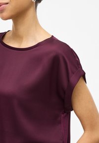 Burgunderfarget kortermet bluse i glatt stoff, med rund hals og rullede ermekanter. Sidespalter for økt bevegelse.