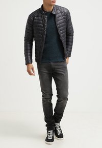 Veste légère matelassée foncée avec un col structuré, associée à un pull vert foncé et un jean gris foncé. Baskets montantes noires.
