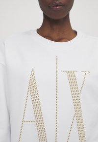 Sudadera blanca con detalles de tachuelas doradas que forman las letras "ALV" en la parte frontal. Textura de tela suave y diseño de cuello redondo.