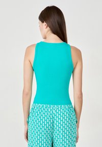 Mujer con cabello largo castaño que lleva una blusa sin mangas de canalé turquesa y pantalones turquesas con patrones florales blancos, vista desde atrás.