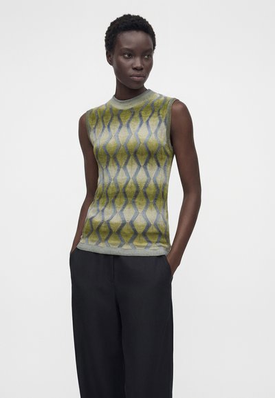 Γυναίκα που φοράει αμάνικη πράσινη και γκρι πουαντεριστή πλεκτή μπλούζα και φαρδιά μαύρη παντελόνα, όρθια με τα χέρια στις τσέπες μπροστά σε λευκό φόντο.