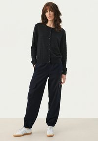 Cardigan negru cu mâneci lungi și nasturi, asortat cu pantaloni cargo bleumarin cu buzunare și adidași albi cu tălpi bej.
