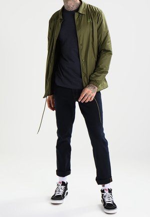 Oliivinvihreä kevyt takki, jossa on kaulus ja nepparit, käytössä tumman t-paidan päällä, yhdistettynä käärittyihin tummiin farkkuihin ja mustiin lenkkareihin.