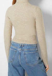 Beige, ribbade turtleneck-tröja med långa ärmar och figurnära siluett, i kombination med högmidjade blå denimjeans som visar bakfickor.