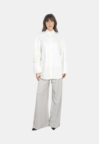 Camicia bianca oversize con bottoni e spalle scese, abbinata a pantaloni larghi grigi chiari e décolleté nere a punta. Tessuto dalla consistenza liscia.