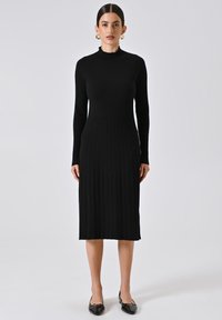 Abito nero a coste con colletto alto, maniche lunghe e silhouette aderente, realizzato in materiale knit elasticizzato. Scarpe nere piatte completano il look.