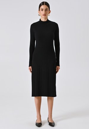 Abito nero a coste con colletto alto, maniche lunghe e silhouette aderente, realizzato in materiale knit elasticizzato. Scarpe nere piatte completano il look.