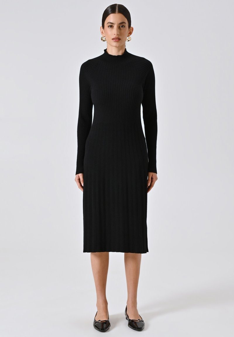 Abito nero a coste con colletto alto, maniche lunghe e silhouette aderente, realizzato in materiale knit elasticizzato. Scarpe nere piatte completano il look.